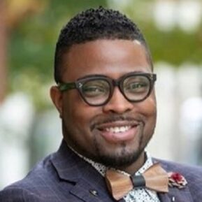 dr o2 (2) Smiling man with short hair, glasses and beard, wearing dark sports jacket, patterned shirt, and gold bow-tie