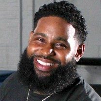 WH Headshot a Black man in his mid-30s with a dark beard and mustache and a smile in his eyes. He is wearing a black sweatshirt and a silver chain around his neck.