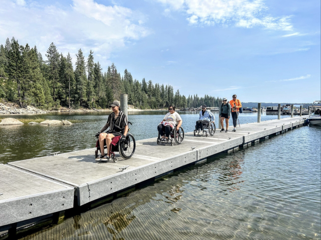 A group of people, including three individuals using wheelchairs and two others walking, move along a floating dock on a calm lake surrounded by pine trees under a sunny, blue sky.